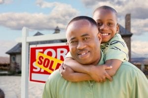 Happy African American Father and Son in Front of New Home and Real Estate Sign.